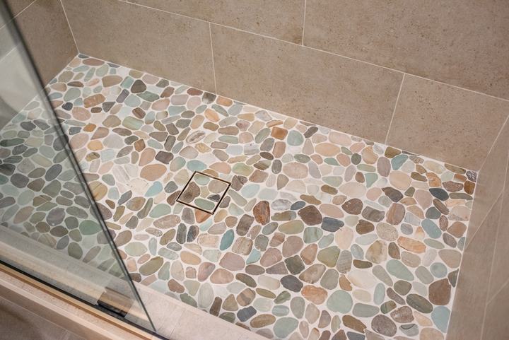 Sliced pebble shower floor with a square tile-in drain, seen past the glass door panel