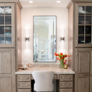 Built-in makeup station flanked by ribbed-glass tower cabinets and an upholstered ring-pull chair.
