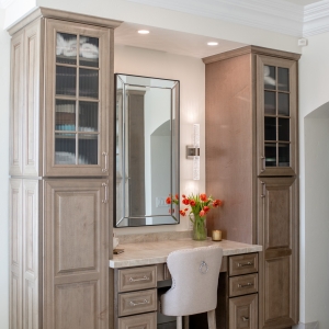 Full-height makeup vanity flanked by ribbed-glass tower cabinets, tulip bouquet and ring-pull chair beneath recessed lights.