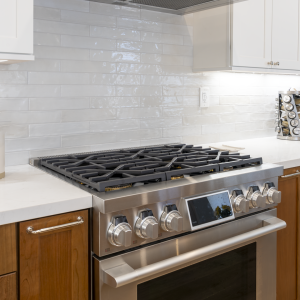 Close-up of stainless professional gas range and hood framed by glossy ceramic backsplash and walnut cabinetry